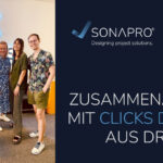 5 Personen (3Männer und 2 Frauen), die lächelnd in die Kamera schauen, sie stehen vor einer weißen Wand mit Leuchtschild "Clicks Digital"; daneben ist ein Rechteck worauf "Zusammenarbeit mit clicks digital aus Dresden" steht, oben ist das SONAPRO und Clicks digital Lofo