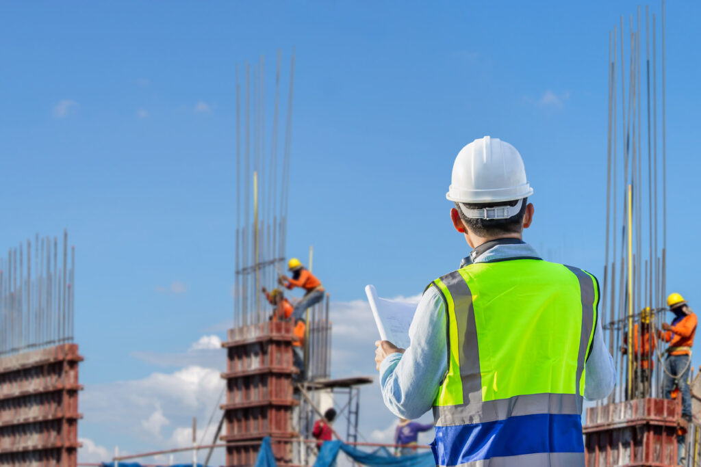 Ingenieur prüft Projektpläne auf einer Baustelle, im Hintergrund unscharf ein Arbeitsteam am Bau